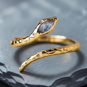 May include: A gold-colored snake ring with a textured finish. The snake's head features a blue-gray stone. The ring is set against a gray, textured surface.