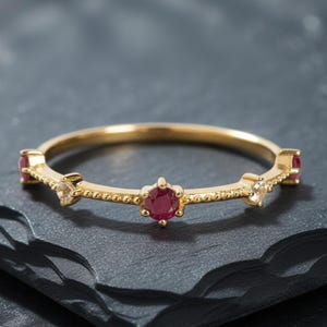 Op de afbeelding: Een gouden bandring met drie rode edelstenen en twee kleinere, heldere edelstenen. De ring heeft een delicaat ontwerp met een getextureerde band. De edelstenen zijn gezet in een klauwzetting. De ring wordt gepresenteerd op een donkergrijze ondergrond.