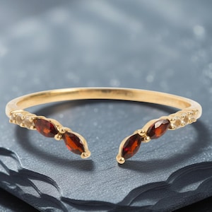 Peut inclure: Une bague dorée ornée de trois pierres précieuses ovales rouge foncé de chaque côté. La bague présente un design à tige fendue, avec les pierres précieuses disposées en motif incurvé. La bague est présentée sur une surface texturée gris foncé.