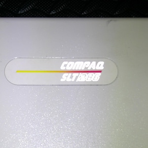 May include: Close-up of a light gray Compaq SLT/286 laptop. The laptop has a white oval sticker with the Compaq logo and SLT/286 model name. The sticker has a yellow, orange, and red stripe.