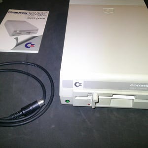 May include: A beige Commodore 1541 disk drive with a front-loading slot, a manual latch lever, and a green power indicator light. Included is a black circular connection cable and a white Commodore 1541C user's guide manual with a blue logo.