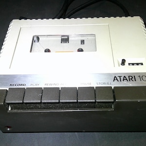May include: An Atari 1010 program recorder featuring a beige top casing with a cassette compartment and a dark gray base. The front panel includes six black rectangular control buttons labeled Record, Play, Rewind, Pause, and Stop/Eject.
