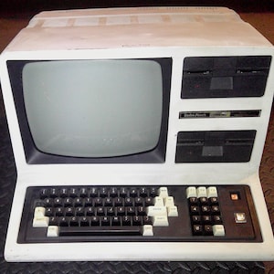 May include: Vintage white computer with a built-in monitor, keyboard, and two floppy disk drives. The keyboard has white keys and a numeric keypad. The computer has the text "Radio Shack TRS-80".