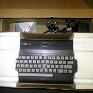 May include: A vintage black computer with a keyboard, nestled in its original cardboard box with styrofoam inserts. The box also contains a power adapter, cables, and a small game cartridge. The computer has a rainbow-colored stripe.