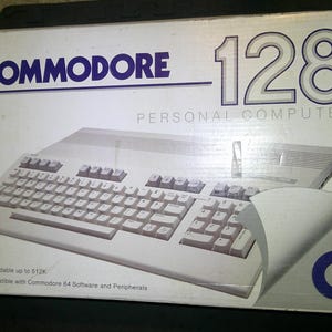 May include: A white cardboard box for a Commodore 128 personal computer. The box features the Commodore 128 logo, a graphic of the computer keyboard, and text stating it is expandable up to 512K and compatible with Commodore 64 software.