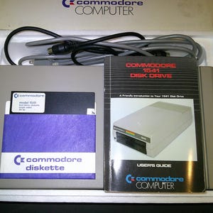 May include: A grey Commodore 1541 disk drive, a black and white user guide, a grey cable, and a Commodore diskette with a purple sleeve, all housed in a white styrofoam packaging box with the text Commodore COMPUTER printed on the top edge.