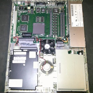 May include: The interior of a beige computer chassis featuring a green motherboard with various integrated circuits, a black cooling fan, a circular speaker, a rectangular hard drive, and a metallic power supply unit with a TDK label.