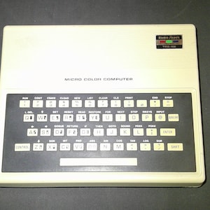 May include: A vintage Radio Shack TRS-80 Micro Color Computer. The off-white computer has a black keyboard with white keys and text. The top right corner has a Radio Shack logo and the text "TRS-80". The text "MICRO COLOR COMPUTER" is printed above the keyboard.