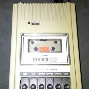 May include: A beige Atari 410 Program Recorder with a clear cassette door, a mechanical tape counter, and six black control buttons labeled Play, Rewind, Advance, Stop/Ej, and Pause. The device features a black power cord extending from the top.