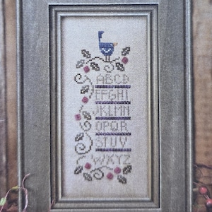 May include: A framed cross-stitch sampler titled "The Plum Berry Sampler" by Bent Creek. The sampler features a blue bird, alphabet letters, and berry sprigs in a vertical design. The frame is a muted gray color, and the background is a wood grain.