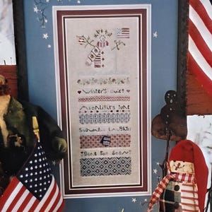 May include: A framed cross-stitch pattern with a patriotic snowman theme. The image includes a framed cross-stitch, a miniature American flag, and a pair of mittens. The text "Patriotic Snowman" is at the top, and "Shepherd's Bush Printworks" is at the bottom.