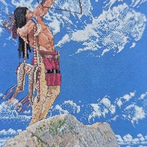 May include: Cross-stitch artwork depicting a Native American figure aiming a bow and arrow. The figure is standing on a rocky outcrop against a blue sky with white clouds. The artwork features detailed stitching and a vibrant color palette.