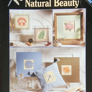 May include: A craft kit titled "Natural Beauty" featuring cross-stitch designs. The image showcases framed needlework of a seashell, fern, rose, daisy, and leaf. A dragonfly pillow is also included. The kit is from Dimensions.