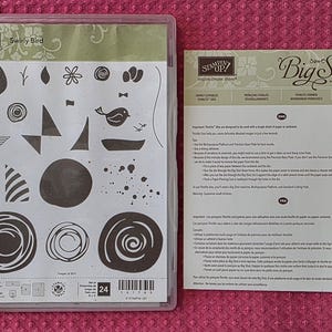 May include: A Stampin' Up! Swirly Bird clear stamp set in a plastic case featuring various bird, floral, and abstract shapes, accompanied by a Big Shot instruction sheet with text in English, French, German, and Japanese detailing usage.