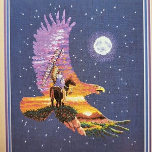 May include: Cross-stitch artwork depicting a Native American on horseback within the silhouette of an eagle. The design features a night sky with stars and a full moon, with the eagle's wings incorporating a sunset landscape. The artwork is framed.
