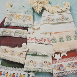 Peut inclure: Une collection de serviettes brod&eacute;es avec des motifs d'anges et de ch&eacute;rubins. Les serviettes pr&eacute;sentent diverses couleurs, dont le blanc, le vert et le bordeaux, avec une bordure en dentelle. Certaines serviettes ont des d&eacute;corations en forme d'&eacute;toile. Le texte "produced by Rita Weiss" est visible.