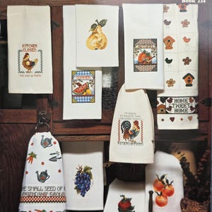 May include: A collection of white Heirloom Huck Towels with various embroidered designs. The designs include a rooster, fruit, vegetables, and text. The towels are displayed on a wooden surface. The text on the towels includes "Kitchen Closed" and "Home Sweet Home".