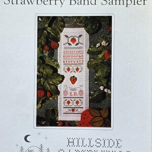 May include: A printed pattern for a cross-stitch sampler titled "Strawberry Band Sampler." The design features a central panel with an alphabet, strawberries, and decorative elements. The image is framed by strawberry plants and foliage.