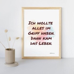 May include: A framed print with the text "ICH WOLLTE ALLES IM GRIFF HABEN. DANN KAM DAS LEBEN." in dark brown lettering over a watercolor background of pink, yellow, and blue hues. The frame is light brown.