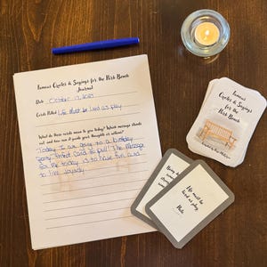 May include: A journal page with handwritten notes and the title "Famous Quotes & Sayings for the Park Bench Journal" is next to a stack of cards with the same title. A lit candle in a glass holder and a blue pen are also on the wooden surface.