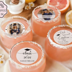 May include: Four stemless glasses filled with a pink beverage, each with a white rim. The glasses feature edible toppers with graduation-themed designs and text. The image includes text that says "100% Edible Toppers Edible Ink".