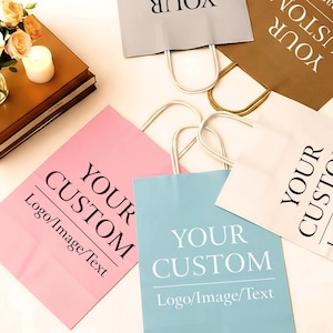 May include: A selection of custom shopping bags in shades of pink, blue, grey, and brown. Each bag displays the text "YOUR CUSTOM" and space for a logo or image. The bags have white and gold handles.