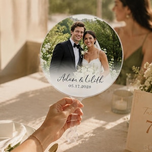 May include: A circular clear acrylic hand fan featuring a printed photograph of a couple in wedding attire. The text "Adam & Lilith" is printed in a black script font above the date "05.17.2028" in a simple sans-serif font on the lower portion.