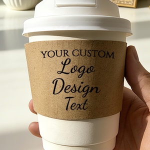 May include: A white paper coffee cup with a white lid and a brown paper sleeve. The sleeve features the text "YOUR CUSTOM Logo Design Text" in black. The cup is held by a hand.