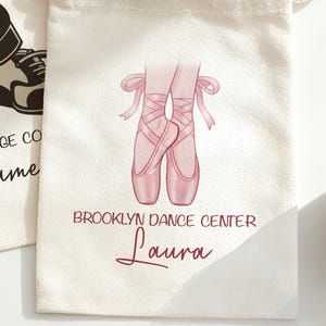 May include: Off-white drawstring bag featuring a watercolor illustration of pink ballet pointe shoes with ribbons. The text "BROOKLYN DANCE CENTER" and the name "Laura" are printed in burgundy.