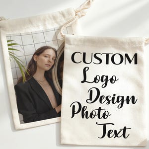 Op de afbeelding: Twee crèmekleurige trekkoordtassen. Eén tas toont een foto van een persoon in een zwarte blazer. De andere tas heeft de woorden "CUSTOM Logo Design Photo Text" in zwart script.