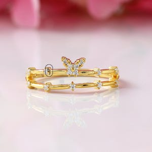 Può includere: Un anello color oro con un design a farfalla e piccole pietre preziose trasparenti. L'anello ha un design a doppia fascia, con la farfalla in cima e pietre preziose lungo le fasce. L'anello è posto su uno sfondo rosa tenue.