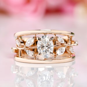 以下が含まれることがあります： オーバルカットのセンターストーンと葉の形をしたアクセントが付いたローズゴールドのリング。リングは、複数の小さな石を備えた繊細なオープンワークデザインが特徴です。リングはソフトピンクの背景にセットされています。