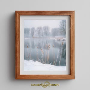 Puede incluir: Una impresión enmarcada de un sereno paisaje invernal. La imagen presenta un lago brumoso con árboles desnudos reflejados en el agua. Juncos altos y secos se alzan en primer plano, con nieve cubriendo el suelo. El marco es de madera marrón claro, con "GOLDEN VALE PRINTS".