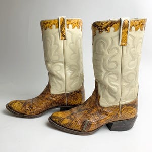May include: A pair of cowboy boots featuring a cream-coloured leather shaft with decorative stitching and a brown snakeskin-textured leather foot. The top edge of the shaft is trimmed with a matching snakeskin pattern and includes pull tabs.