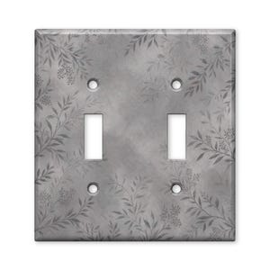 May include: A rectangular double light switch wall plate featuring a gray background with a subtle, darker gray botanical pattern of delicate leaves and small berries. The plate includes two vertical rectangular cutouts and four screw holes.