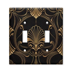 May include: A double light switch wall plate featuring a black background with a symmetrical gold filigree pattern. The design includes stylized fan-shaped motifs, curved lines, and small dots. It has two rectangular cutouts and four screw holes.