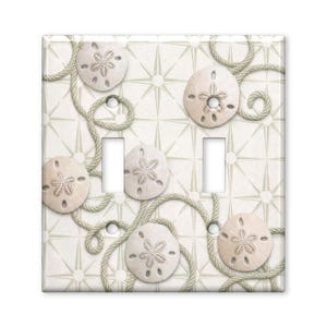 May include: A double light switch wall plate featuring a light beige background with a repeating star pattern. The design includes several off-white sand dollars and coiled light tan rope accents arranged across the rectangular surface.
