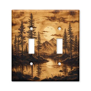 May include: A double light switch plate featuring a wood grain finish with a dark brown engraved landscape scene. The design depicts a serene lake surrounded by tall pine trees and jagged mountains under a large full moon with birds flying.