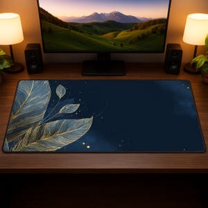 May include: A navy blue desk mat featuring a gold feather and leaf design. The mat is rectangular and sits on a wooden desk. A computer monitor, speakers, and lamps are visible in the background.
