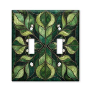 May include: A double toggle light switch plate cover featuring a symmetrical stained glass pattern of overlapping dark and light green leaves with thin dark outlines. The rectangular plate has two vertical rectangular cutouts for switches.