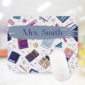 May include: A personalized mousepad featuring a white background with colorful illustrations of calculators, rulers, and other school supplies. The name "Mrs. Smith" is displayed in a blue banner. A white computer mouse is next to the mousepad.
