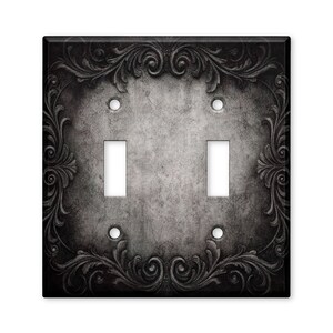 May include: A double toggle light switch plate featuring a dark gray and black distressed concrete texture background. The rectangular plate is framed by an ornate, embossed black scrollwork border with intricate swirling leaf patterns.