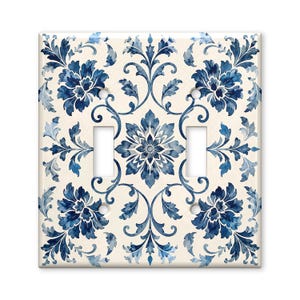 May include: A square double light switch wall plate featuring a cream-colored background with a symmetrical blue floral and leafy vine pattern. The design includes a central star-shaped flower motif and two vertical rectangular cutouts for switches.