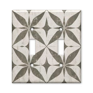 May include: A double light switch wall plate featuring a repeating geometric pattern of dark gray, leaf-shaped motifs on a distressed, off-white background. The rectangular cover includes two vertical rectangular cutouts and four screw holes.