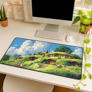 May include: A rectangular desk mat featuring a vibrant illustration of hobbit homes nestled in green hills under a blue sky with fluffy white clouds. The mat has a black border and is designed for use with a computer.