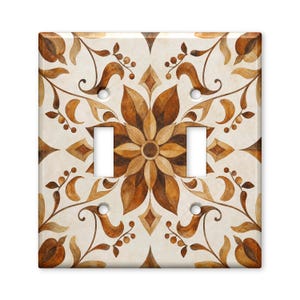 May include: A square double light switch wall plate featuring a symmetrical floral pattern in shades of brown, tan, and cream. The design includes stylized leaves, vines, and berries arranged around a central flower motif with two rectangular cutouts.