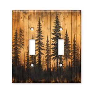May include: A double light switch plate cover featuring a printed design of black pine tree silhouettes against a background of vertical light brown wooden planks. The rectangular plate includes two vertical rectangular cutouts and four screw holes.