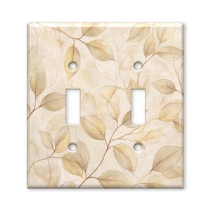 May include: A double light switch wall plate featuring a soft, watercolor-style pattern of overlapping beige and light brown leaves on a cream background. The rectangular cover includes two vertical toggle switch cutouts and four screw holes.