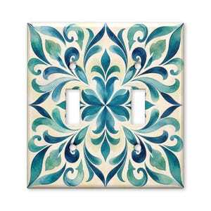 May include: A double light switch wall plate featuring a symmetrical watercolor pattern in shades of teal, navy blue, and cream. The square cover includes two rectangular cutouts for toggle switches and four small white screw head covers.
