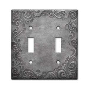 May include: A double toggle light switch plate with a brushed metallic gray finish. The rectangular cover features an embossed scrollwork pattern along the edges and four circular screw holes, with two vertical rectangular cutouts for switches.